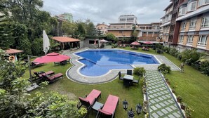 A swimming pool with surrounding lounge chairs and umbrellas, a paved walkway, and a grassy area with potted plants. - The Malla Hotel (Kathmandu)