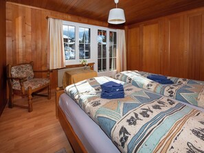 Quarto com painéis de madeira, uma cama, uma cadeira e uma janela com vista para montanhas cobertas de neve.