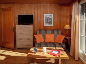 Quarto com armário de madeira, televisão, quadro emoldurado, sofá com almofadas laranja, mesa com toalha de mesa e luminária.