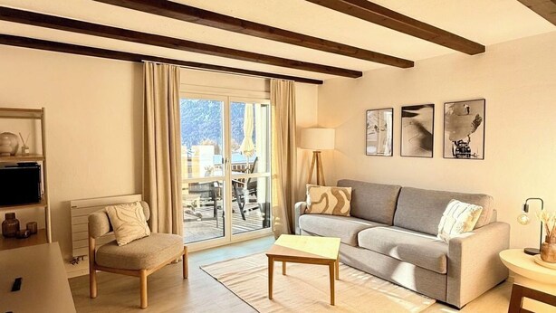 A modern living room with a sofa, a dining table with chairs, a coffee table, and a TV. There is a large window with curtains, a lamp, and artwork on the wall. - Residenza Chesa Margun 58-5 by Interhome (Silvaplana-Surlej)
