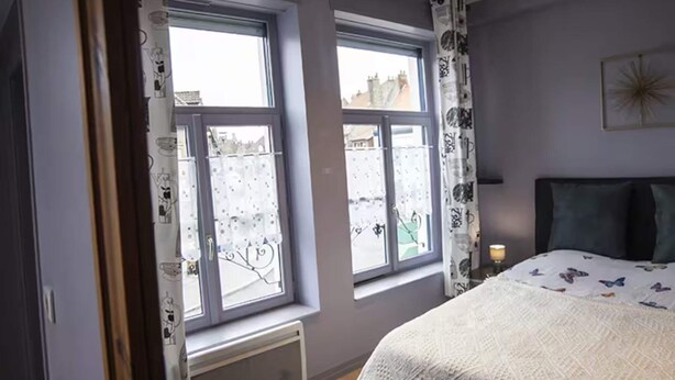 Ein Schlafzimmer mit einem großen Bett, zwei Fenstern mit Vorhängen, einem Heizkörper und Blick nach draußen.