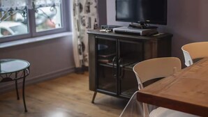 Ein Zimmer mit Holzboden, einem Esstisch mit Stühlen, einem Fernseher auf einer Kommode und einem Fenster mit Vorhängen.