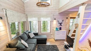 Hyggelig interiør i en hytte med en hjørnesofa, et lille hvidt tekøkken og et stort vindue med udsigt til skoven.