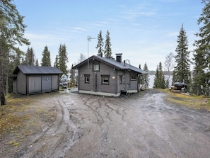 A two-story cabin with a garage, surrounded by trees and a gravel driveway.