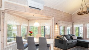En rummelig opholdsstue med spisebord, grå stole, en grå sofa og et stort vindue med udsigt over grøn vegetation.