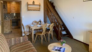 Un salon chaleureux avec un coin repas, un escalier en bois et une cuisine visible à travers une porte ouverte.