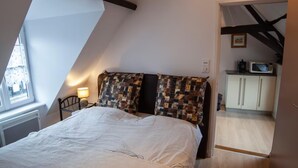 Un dormitorio con una cama grande, una mesita de noche, una lámpara, un cabecero acolchado y vistas a una cocina a través de una puerta abierta.