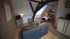 Una habitación estilo loft acogedora, con techo alto, vigas de madera a la vista, un sofá con cojines estampados, una pequeña mesa de comedor con sillas y un televisor sobre un mueble blanco.