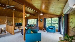 Ein großzügiger Wohnbereich mit Holzboden, einem Fernseher, einer Couch, einem blauen Sessel und großen Fenstern mit Blick auf Grünflächen.
