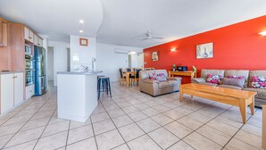A modern living room with a red accent wall, a central island with bar stools, a beige sofa, and a wooden coffee table. - Ocean Views Galore - Airlie Beach (Cannonvale)