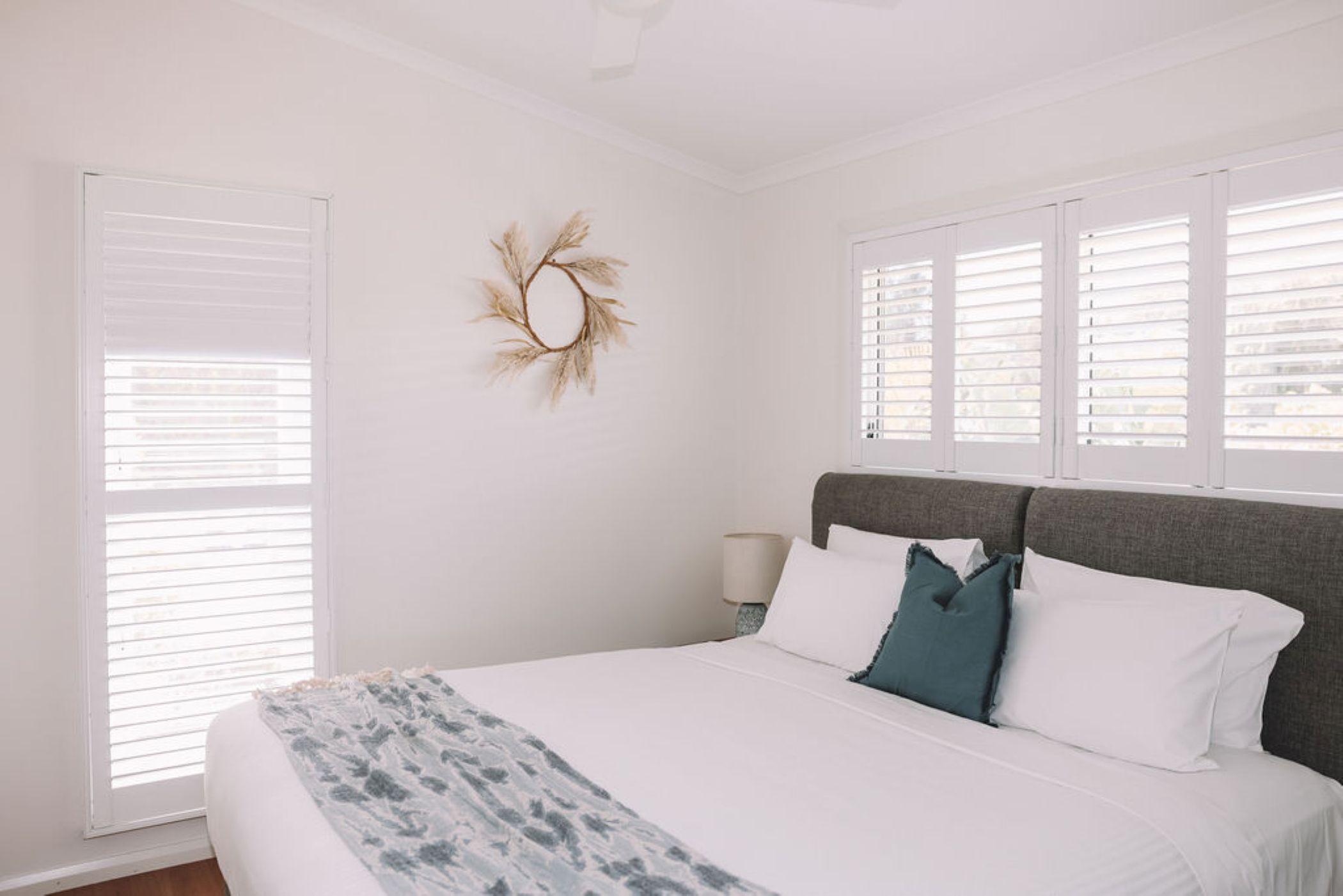 A bedroom with a bed, white walls, a decorative wreath, and a window with blinds.