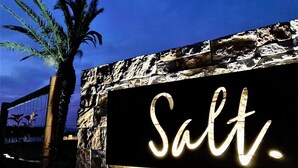A sign with the word "Salt" illuminated in white against a dark background, mounted on a stone wall with a palm tree to the left.