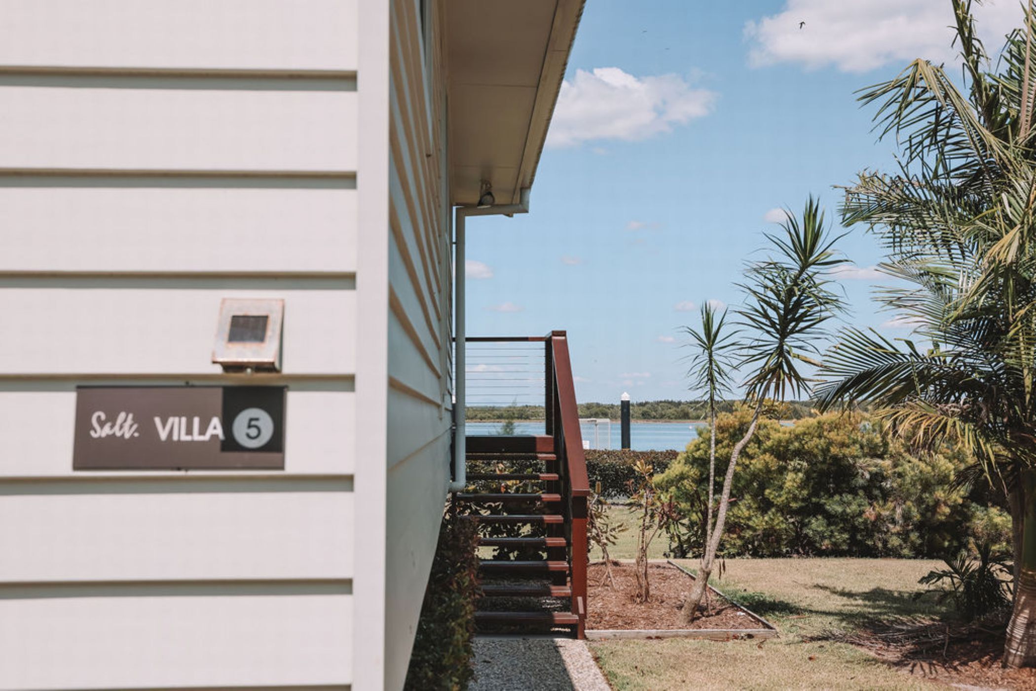 A building with a sign reading "Salt. VILLA 5" and a staircase leading to a balcony overlooking a body of water and palm trees.