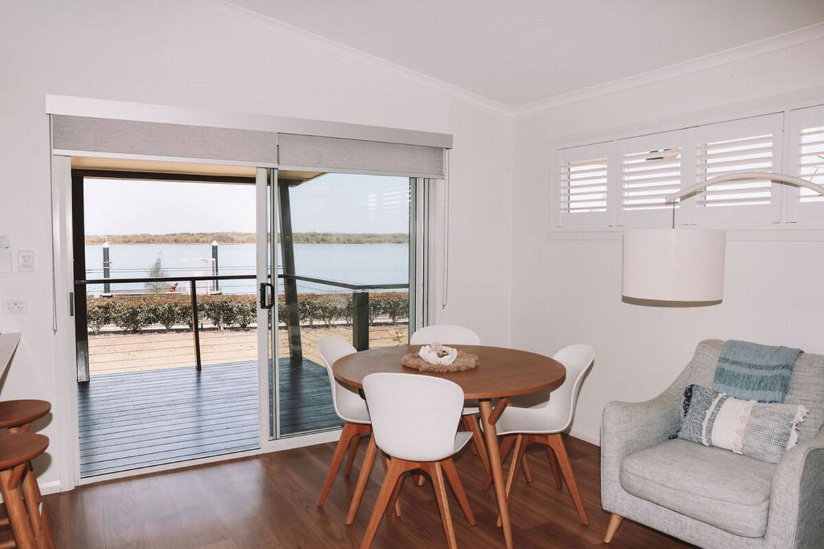 Ein modernes Wohnzimmer mit Essbereich, große Schiebeglasüren zum Deck mit Blick auf ein Gewässer und eine entfernte Landschaft.