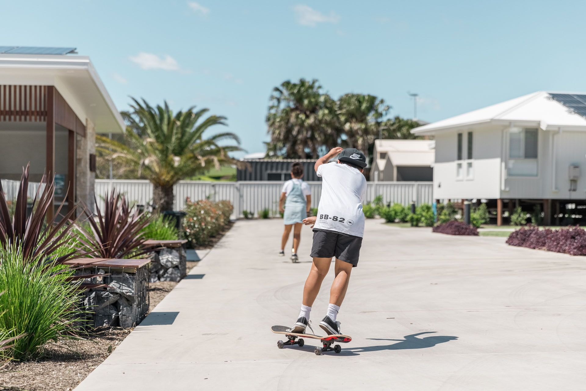 Une personne fait du skateboard sur une allée pavée dans un quartier résidentiel, avec des maisons et des palmiers.