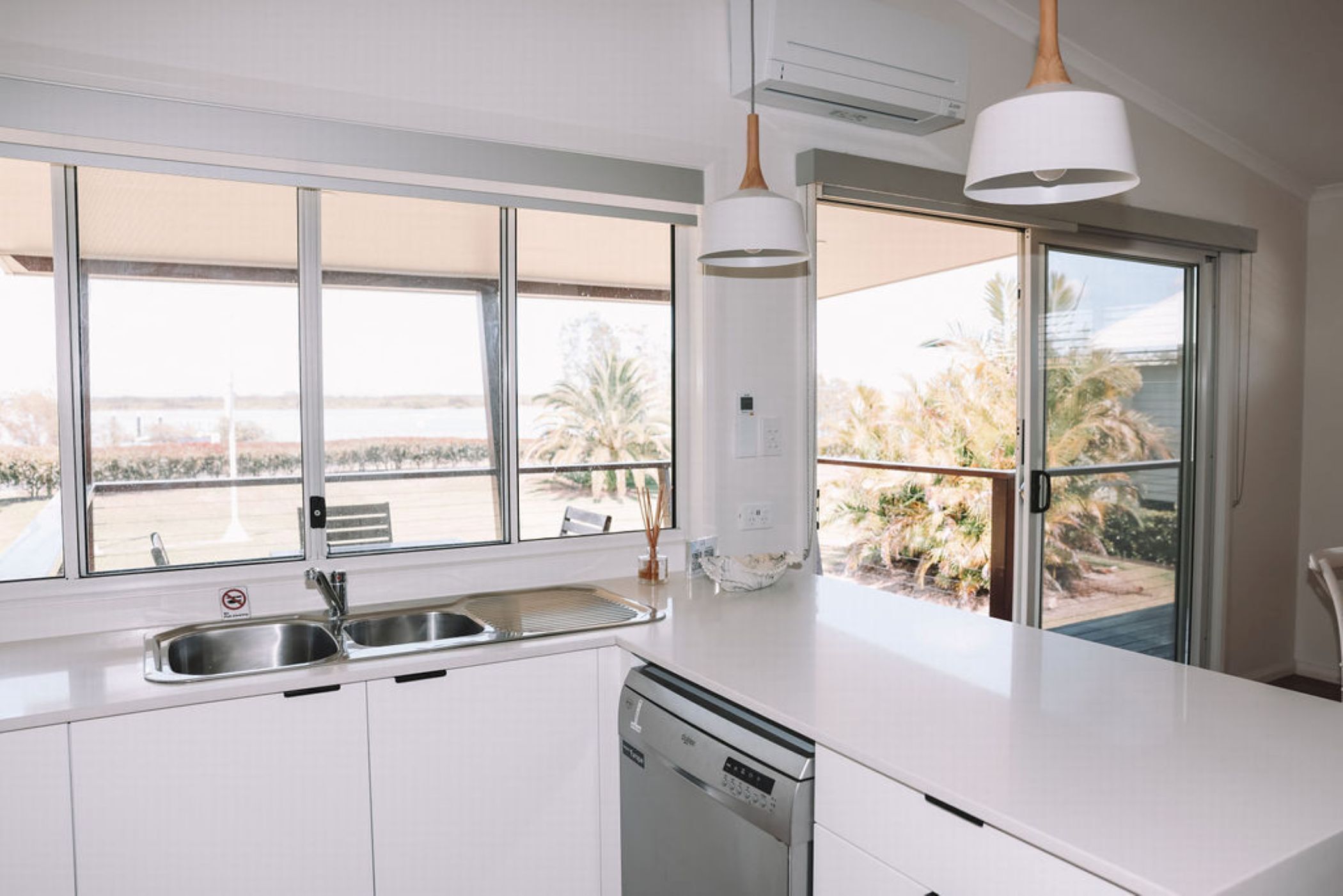 A modern kitchen with a dishwasher, a sink, and a window overlooking a balcony with palm trees.