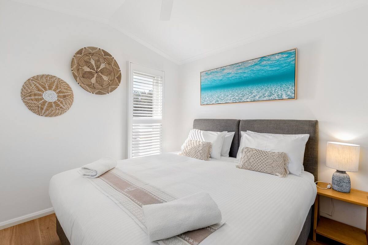 Ein Schlafzimmer mit einem Bett, zwei Wanddekorationen, einem Fenster mit Jalousien, einem Strandbild und einer Nachttischlampe.