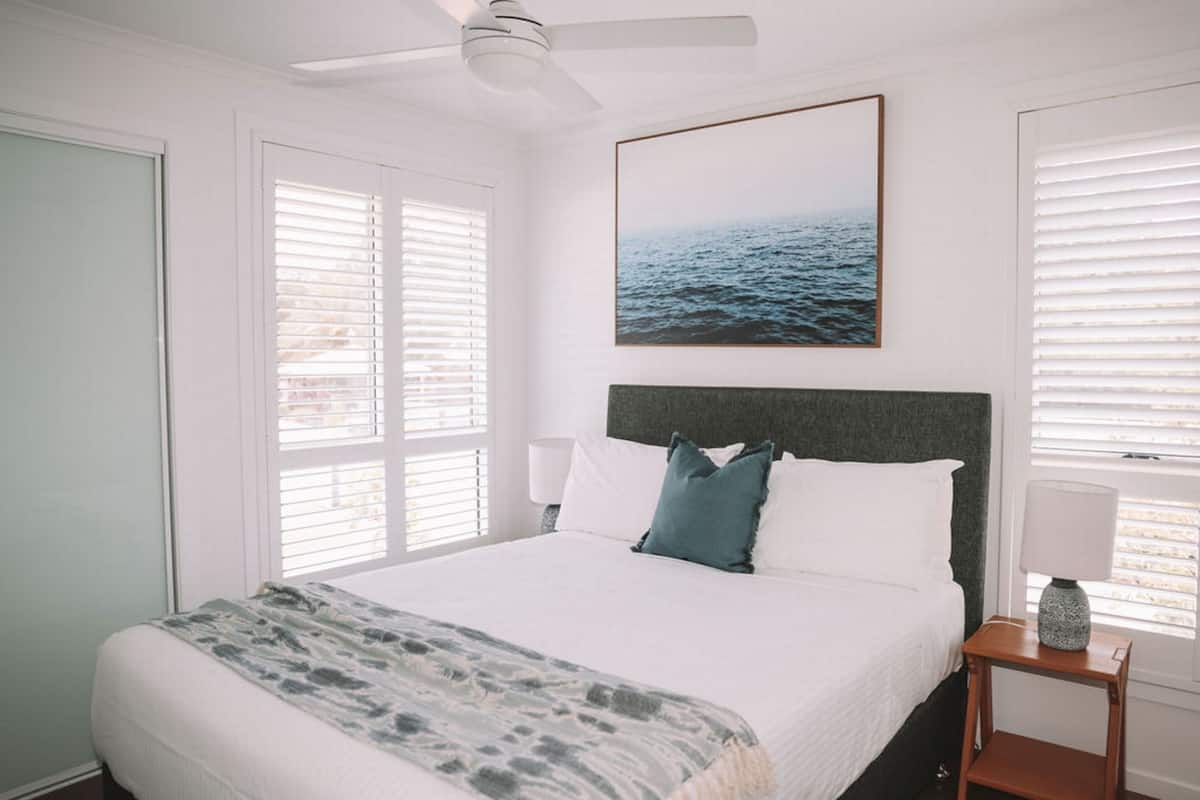 Ein Schlafzimmer mit einem Bett, einem Deckenventilator, einem gerahmten Bild an der Wand und einem Fenster mit Jalousien.