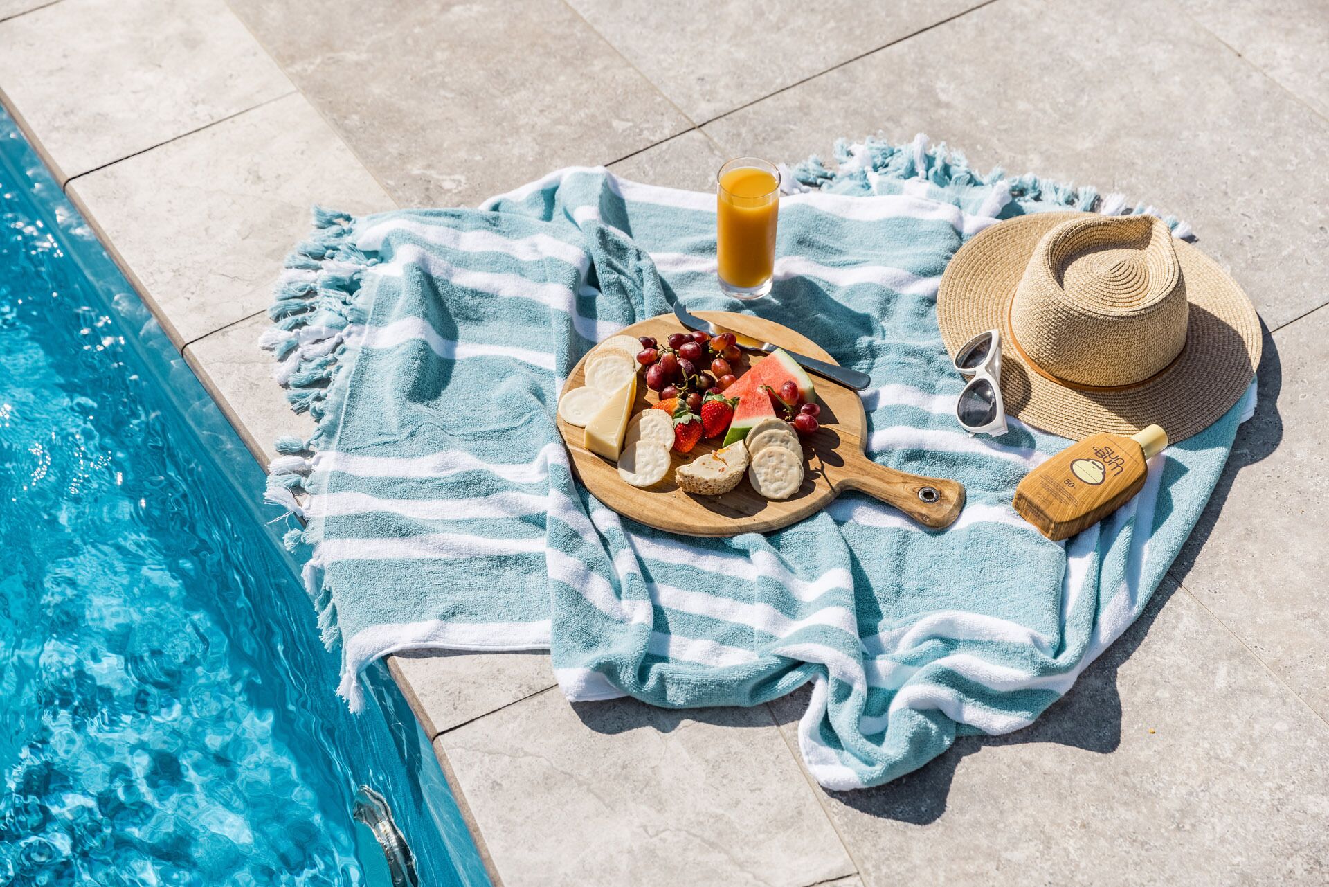 Preparación junto a la piscina con una toalla, una tabla de madera con queso, uvas, fresas y sandía, un vaso de zumo de naranja, gafas de sol y un sombrero.