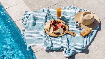 Preparación junto a la piscina con una toalla, una tabla de madera con queso, uvas, fresas y sandía, un vaso de zumo de naranja, gafas de sol y un sombrero.