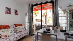 A room with a sofa, dining table, chairs, and a view of palm trees and buildings. - Parque Santiago II 228 - Two Bed (Playa de la Américas)
