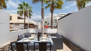 An outdoor dining area with tables and chairs, palm trees, and buildings in the background. - Parque Santiago II 228 - Two Bed (Playa de la Américas)
