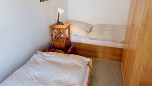 A small, compact room with two bunk beds, a wooden side table with a lamp, and a colorful abstract painting on the wall.