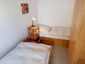 A small, compact room with two bunk beds, a wooden side table with a lamp, and a colorful abstract painting on the wall.