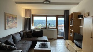 A living room with a sofa, coffee table, TV stand, and a view of a forested area through large windows.