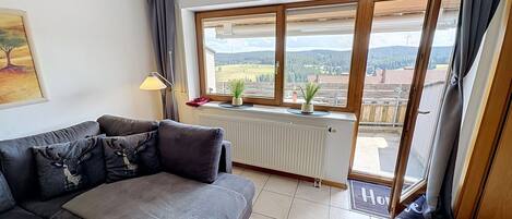 Una sala de estar con un sofá, un radiador, un ventanal con vistas al campo y una puerta que da al exterior.