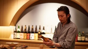 A person holding a bottle in a well-lit room with a wooden counter and various bottles in the background. - KAMAKURA Hotel (Kamakura)