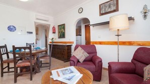 Een woonkamer met een bank, fauteuil, eettafel en stoelen. Er is een lamp, een klok en een schilderij aan de muur.
