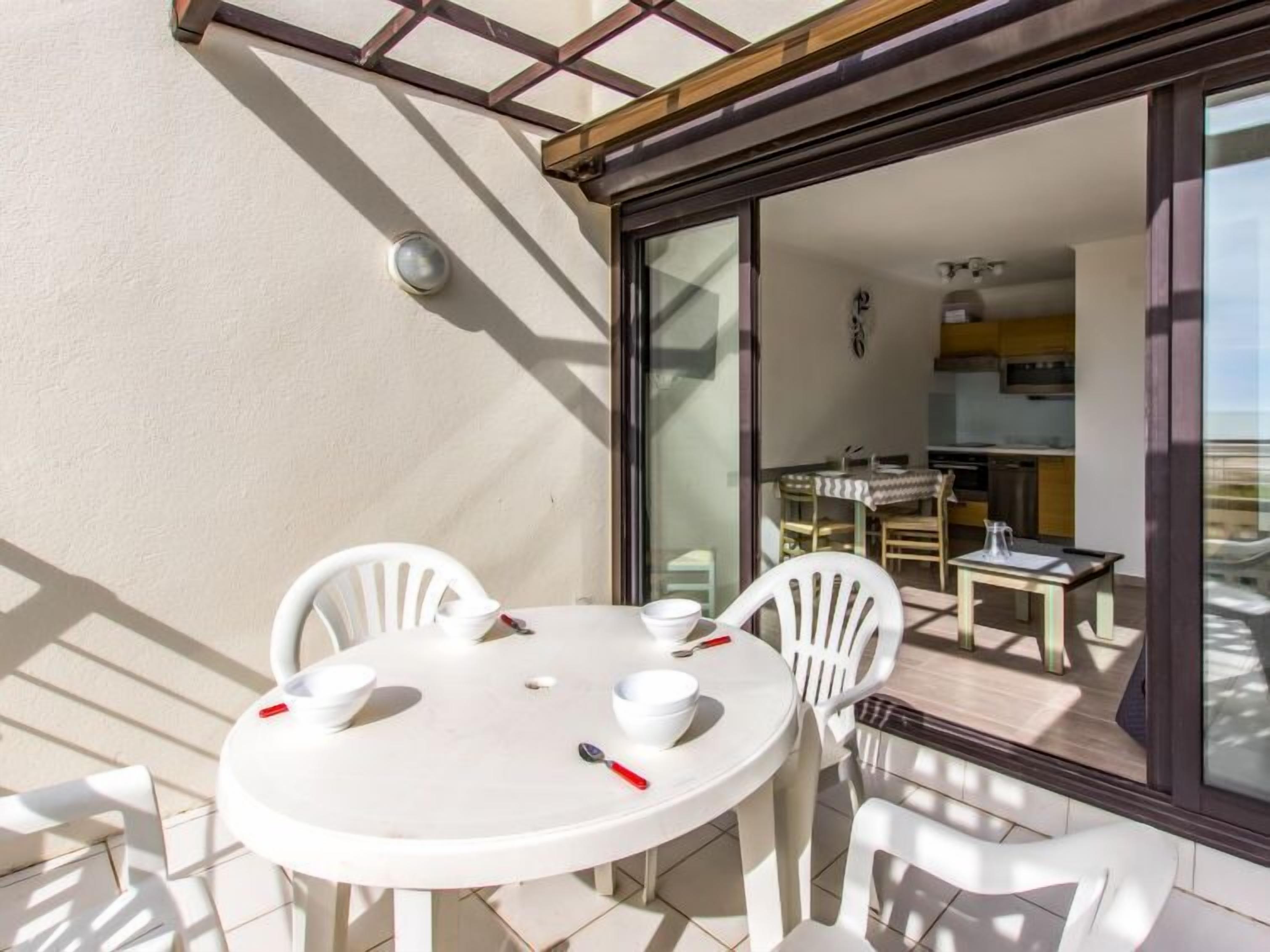 Een zonnig terras met een ronde tafel en vier witte stoelen, een glazen schuifdeur naar een keuken en een klein zitgedeelte buiten.
