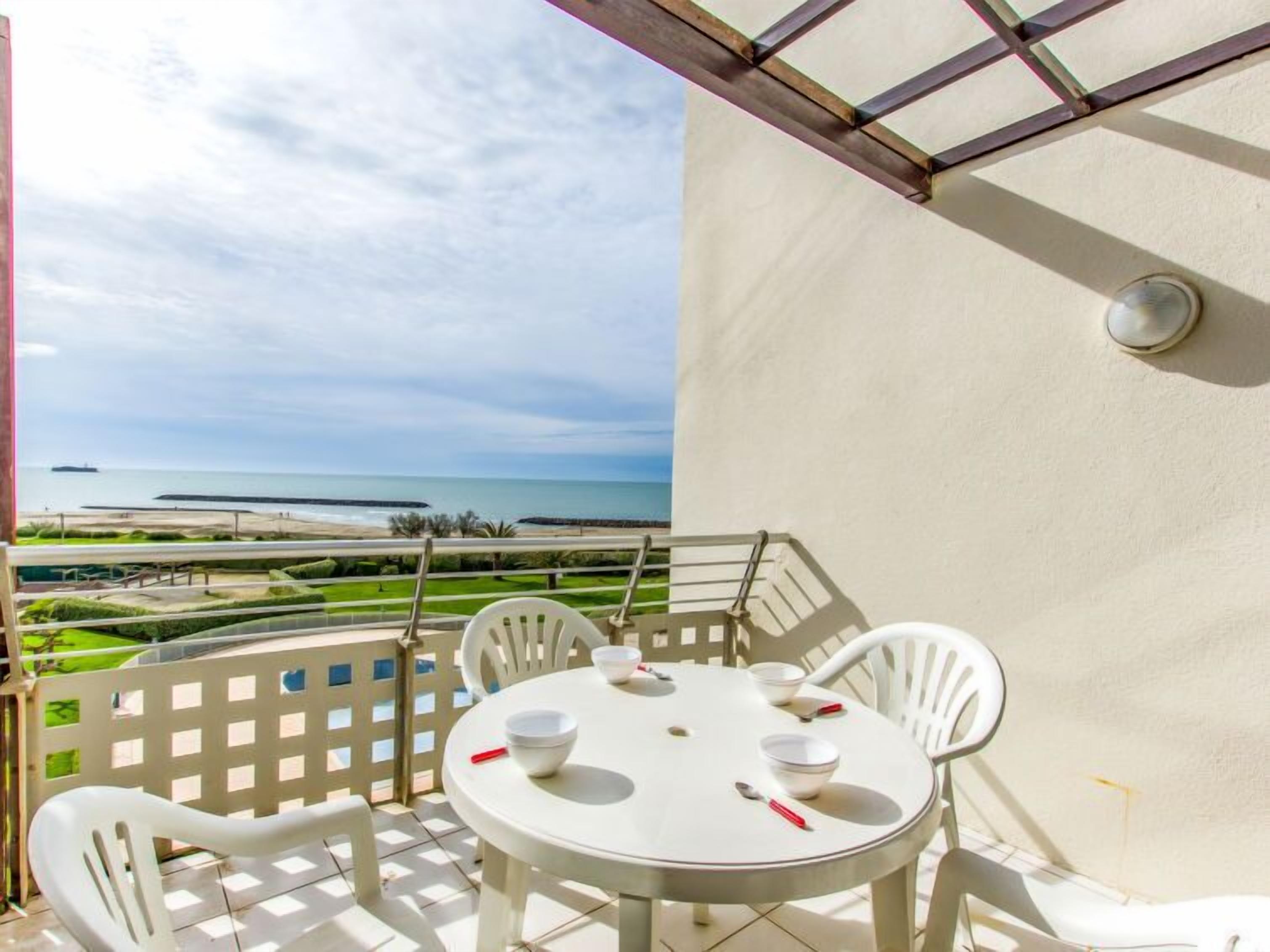 A balcony with a white table and chairs, overlooking a beach and ocean.