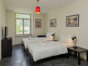 Ein Hotelzimmer mit zwei Betten, einem Fenster mit Blick auf Gebäude, einer roten Pendelleuchte und drei gerahmten Bildern an der Wand.