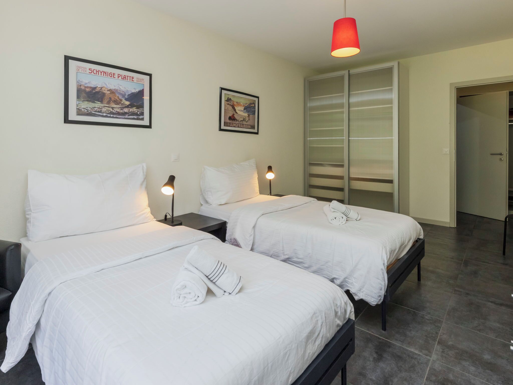 Ein Hotelzimmer mit zwei Betten, einer roten Pendelleuchte und zwei gerahmten Postern an der Wand.