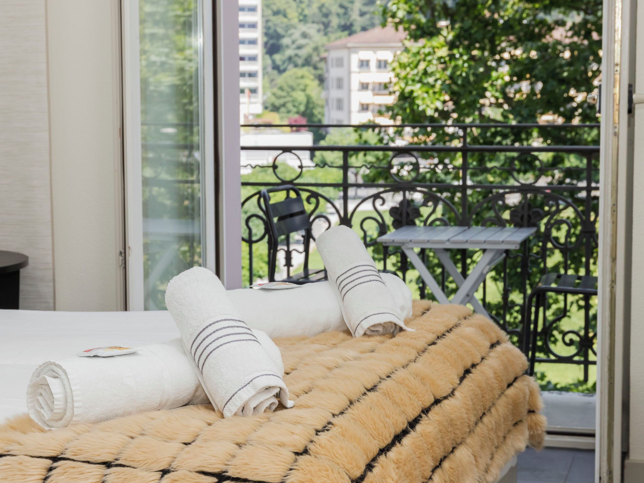 Ein Hotelzimmer mit Bett, einem Fenster mit Blick auf Grünflächen und Gebäude sowie einem Balkon mit Tisch und Stuhl.