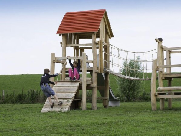 A wooden play structure with a slide and climbing net, set in a grassy field.