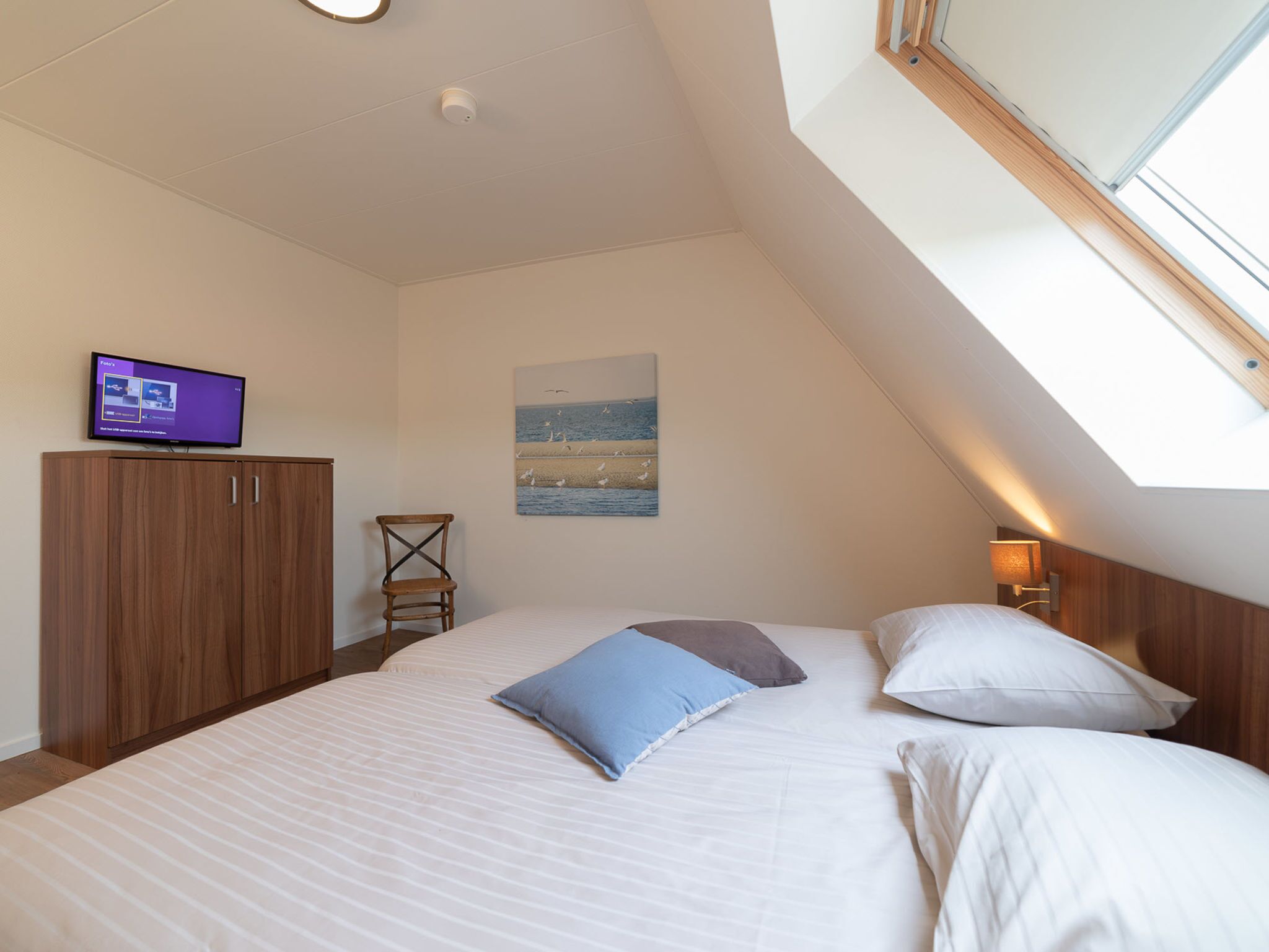 Une chambre à coucher avec un lit, un téléviseur posé sur un meuble, une chaise et un tableau accroché au mur.