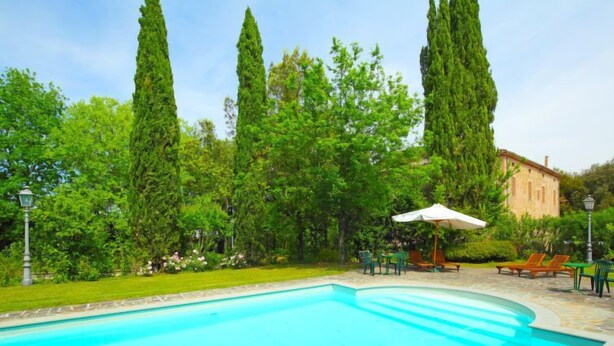 A swimming pool with a stone border, surrounded by lush greenery and tall trees. There are outdoor seating areas with umbrellas and chairs. - Montecorneo by Interhome (Perugia)
