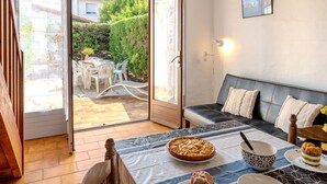 Une pièce chaleureuse avec un canapé, une table à manger sur laquelle se trouvent un gâteau et un saladier, et une vue sur un jardin, visible à travers une porte vitrée.