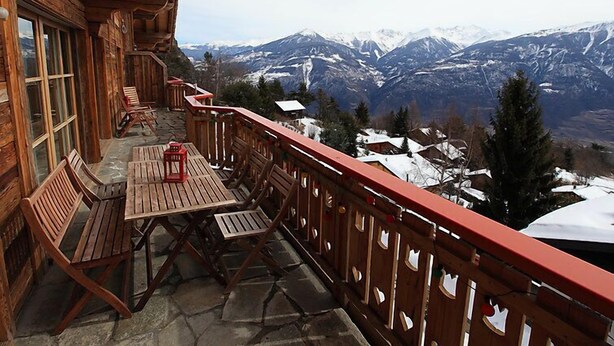 Un balcon en bois, agrémenté d’une table et de chaises, qui offre une vue sur un paysage montagneux aux sommets enneigés.