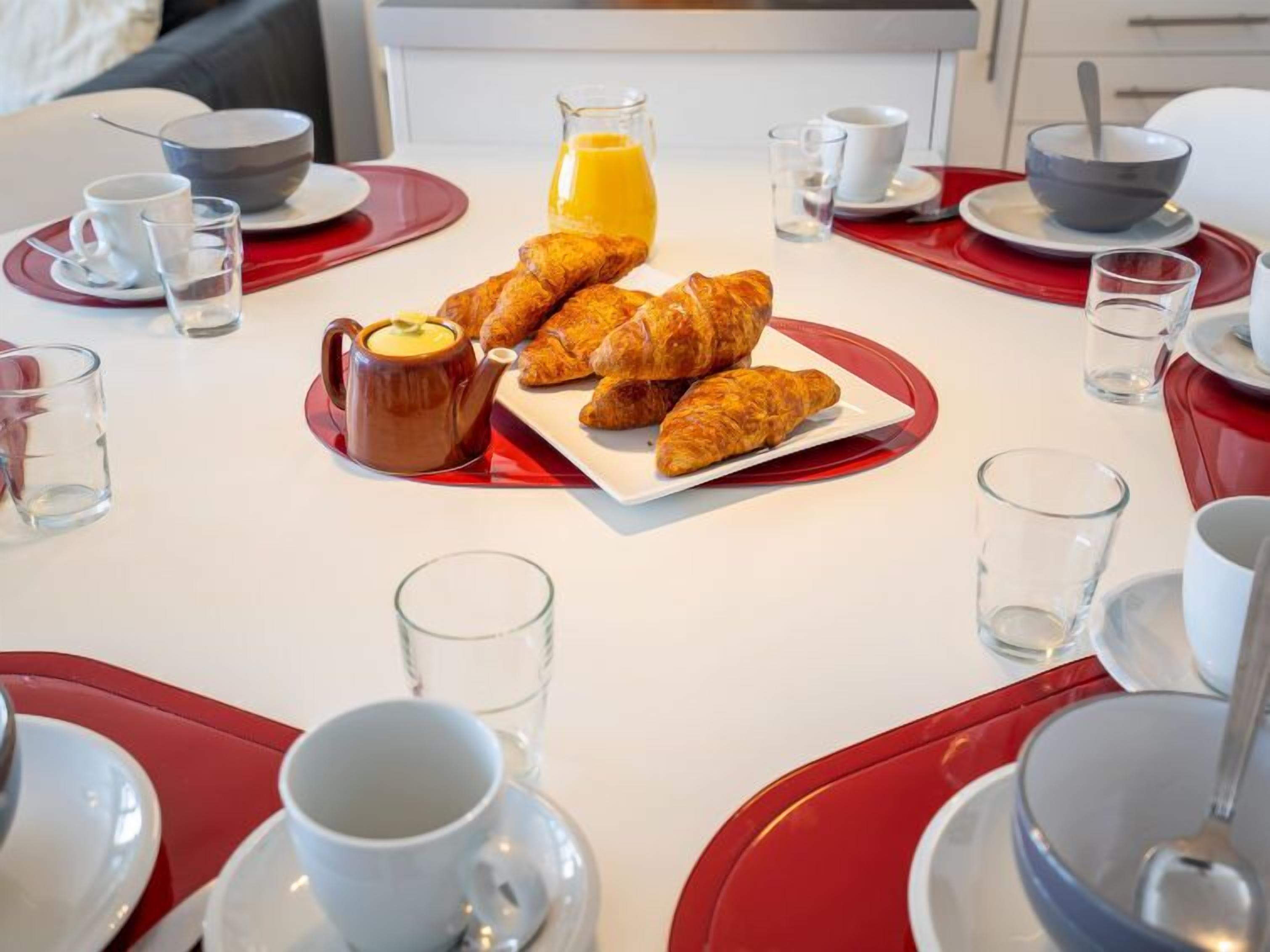 A breakfast setting with croissants, a jug of orange juice, and various cups and bowls on a table.
