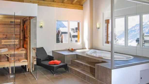 A modern bathroom with a large bathtub, a wooden bench, and a glass shower enclosure. - Apartment A01 by Interhome (Leukerbad)