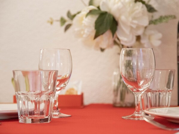 Una mesa puesta con cristalería transparente y platos, un mantel rojo y un jarrón con flores blancas al fondo.