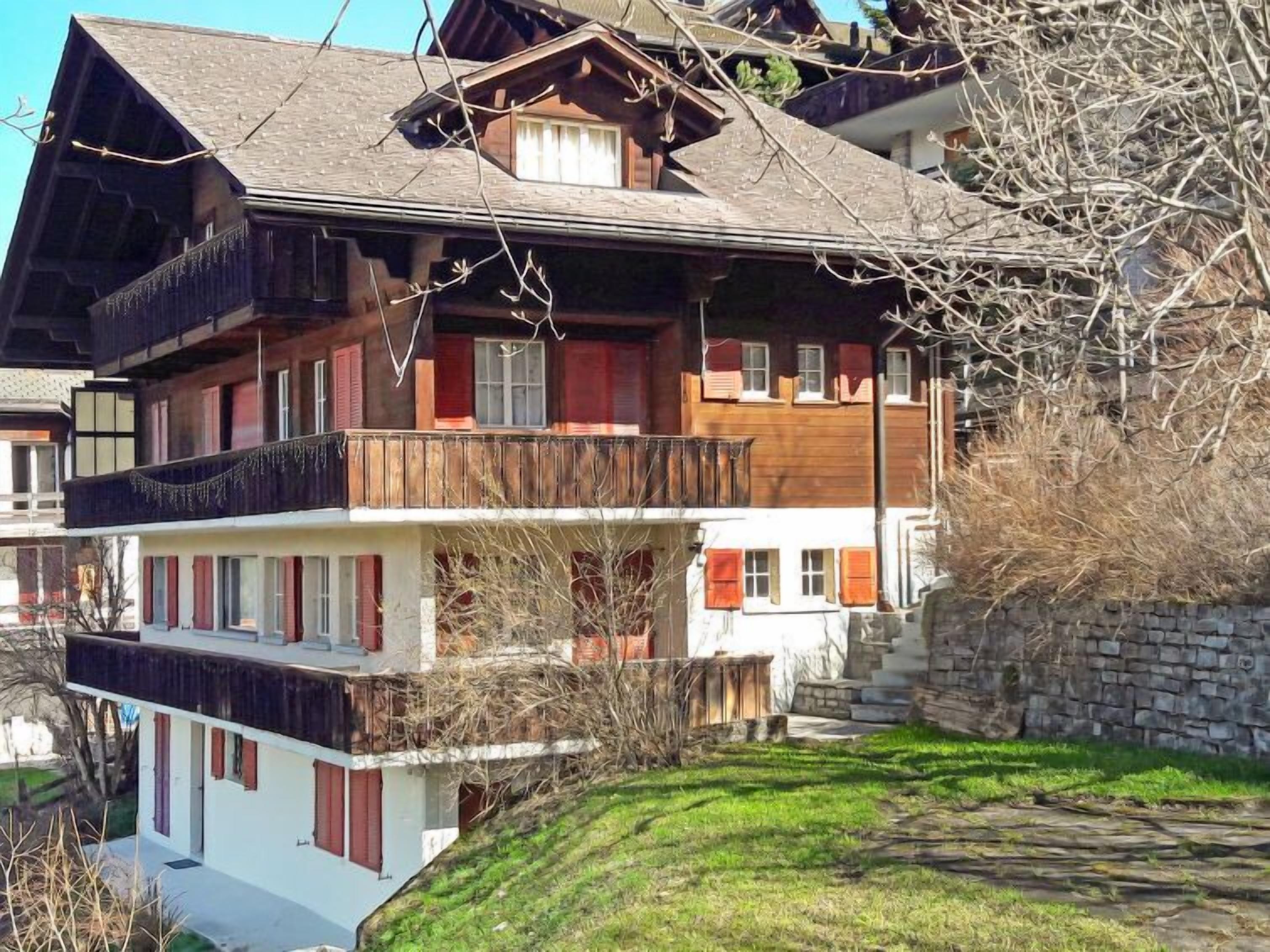 A multi-story chalet with balconies, a stone wall, and a grassy yard.