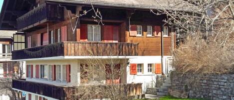 A multi-story chalet with balconies, a stone wall, and a grassy yard.