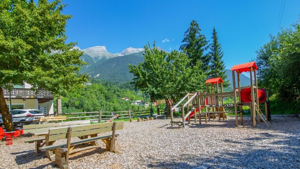 A playground with wooden benches and a red slide, surrounded by trees and mountains in the background. - Kaufmann by Interhome (Pians)