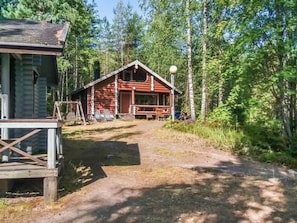 Een chalet met een veranda, een pad van aarde dat ernaartoe leidt, en een bebost landschap op de achtergrond.