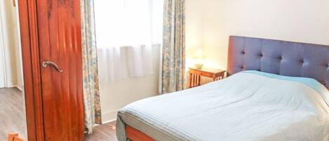 A bedroom with a bed, a wooden wardrobe, a small bedside table with a lamp, and a framed picture on the wall.